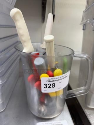 Plastic pitcher containing assorted scoops, spreaders, and bread knives with colored handles. Auction label reads "328".