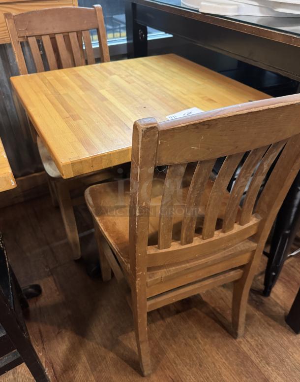 Wooden dining table with two matching chairs, showing wear. Square tabletop with a light finish. Vintage appearance.