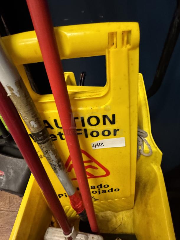 Yellow caution wet floor sign in a cleaning cart with mops. Sign displays "Caution Wet Floor" in English and Spanish. Item tag "442".