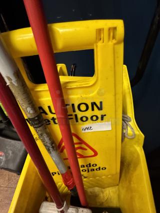 Yellow caution wet floor sign in a cleaning cart with mops. Sign displays "Caution Wet Floor" in English and Spanish. Item tag "442".
