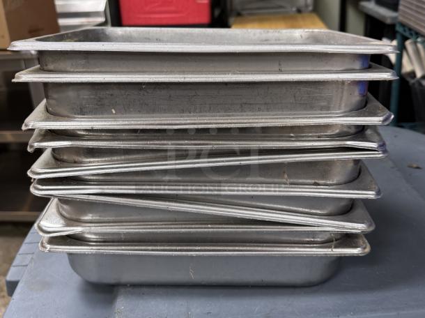 Stack of nine stainless steel half-size pans, used condition, slight wear visible. Ideal for commercial kitchen use.