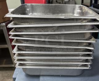 Nine stainless steel half pans stacked. Used condition with visible wear. Ideal for food service or catering.