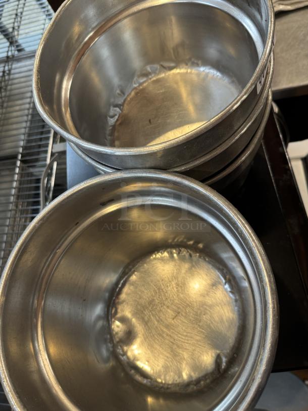 Assorted sizes of round stainless steel insert pans, visibly stacked, in used condition. Four pans shown for auction.