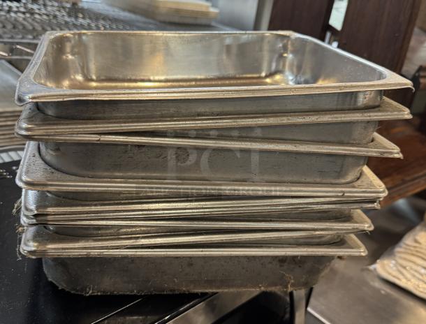 Nine stainless steel half-size sheet pans stacked together, showing visible wear and scratches, suitable for commercial use.