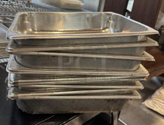 Nine stainless steel half-size sheet pans stacked together, showing visible wear and scratches, suitable for commercial use.