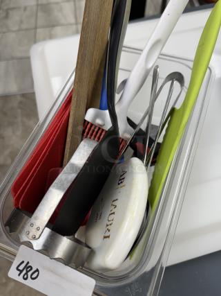 Assorted kitchen items in a clear container, including a wooden spatula, whisk, peeler, and plastic scraper. Visible tag: "480".