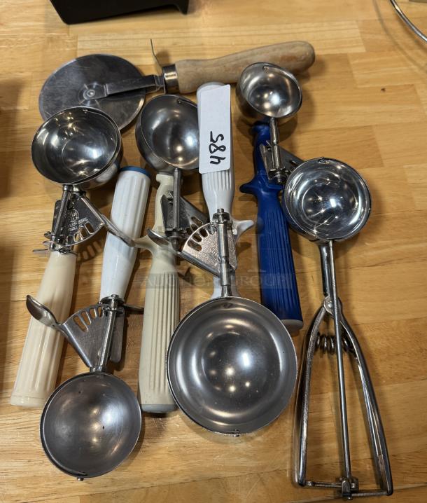 Serving scoops and pizza slicer set on wooden surface, including seven scoops with varied handles and a stainless pizza cutter. Tag labeled "485."