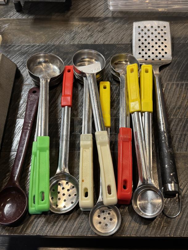 Assorted serving ladles with colored handles and one perforated spatula placed on a wooden surface, showing varying sizes and conditions.