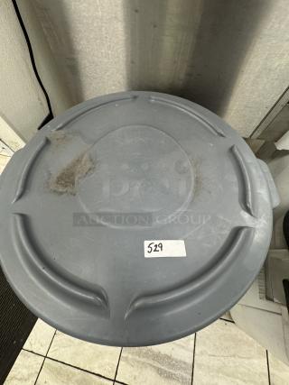 Gray Rubbermaid trash can with lid, used condition, visible label with number 529, slight scuffs and dirt marks.