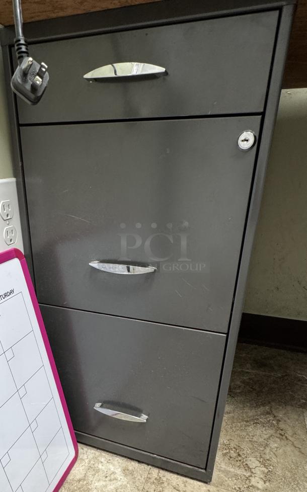 Black filing cabinet under desk, three drawers, metal handles, lock on top drawer, shows minor wear, part of office set.