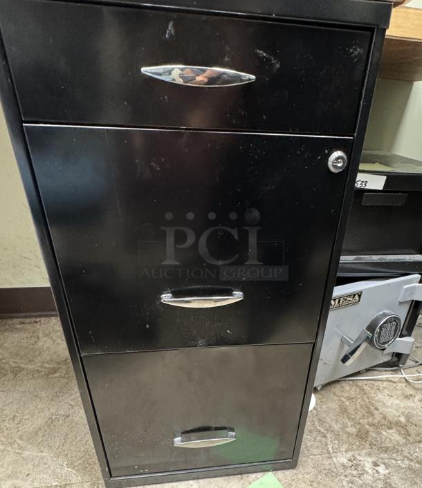 Black filing cabinet with three drawers, silver handles, and a key lock, part of desk set, shows minor wear.