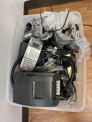 Clear plastic bin containing assorted cables, a Verifone terminal, a receipt printer, and various chargers and cords. Mixed condition.