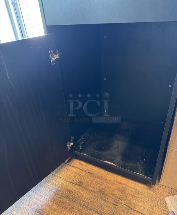Black trash can bin inside a cabinet. Hinged door open, showing interior. Appears functional with minor wear.