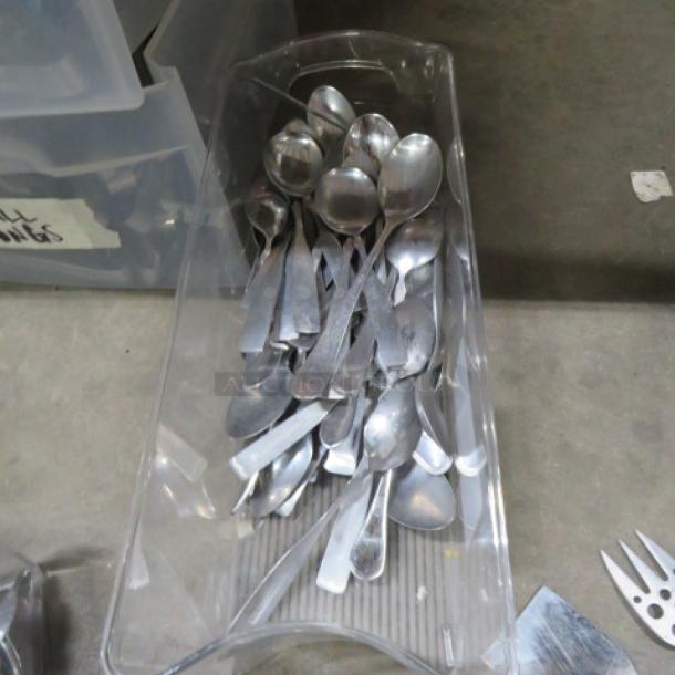 A clear plastic container holds a lot of assorted silver spoons, various sizes, on a concrete surface.