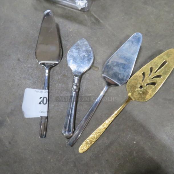 Four assorted pie servers in mixed metals, featuring various designs. Includes a gold-toned server with cut-out pattern. Auction tag visible.