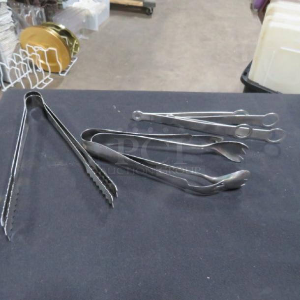 Assorted stainless steel tongs, various types and sizes, in good condition on a black table.