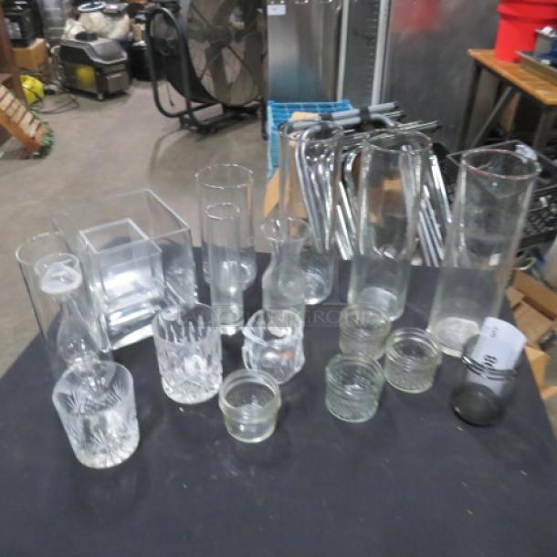 Assorted glassware lot on black table, featuring various shapes and sizes; includes vases, jars, cups, and a clear square dish.
