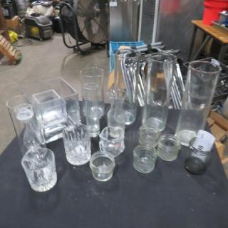 Assorted glassware lot on black table, featuring various shapes and sizes; includes vases, jars, cups, and a clear square dish.