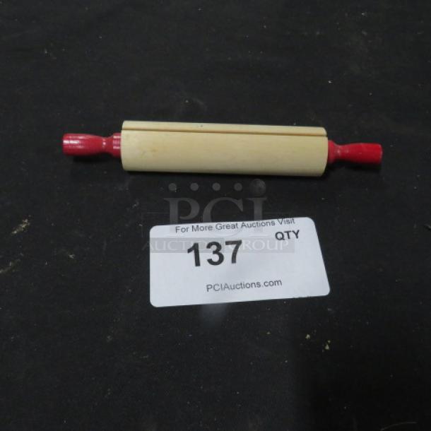 Wooden rolling pin with red handles on a black surface. Auction label "137 QTY" visible.