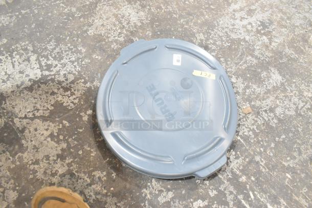 Blue Brute trash can lid, marked "PCL," lays on a textured floor. Appears in good condition, no visible defects.