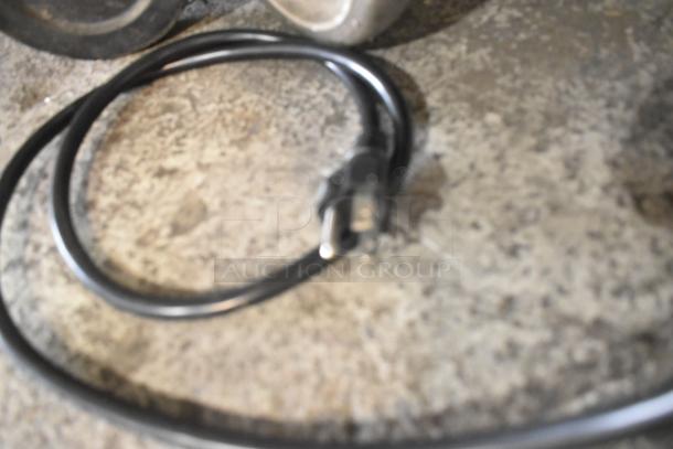 A power cord is coiled on a textured surface, connected to the Vollrath TW-27R food warmer, indicating its electrical functionality.