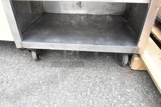 Stainless steel counter with under shelves, featuring caster wheels for mobility, in used condition on pavement.