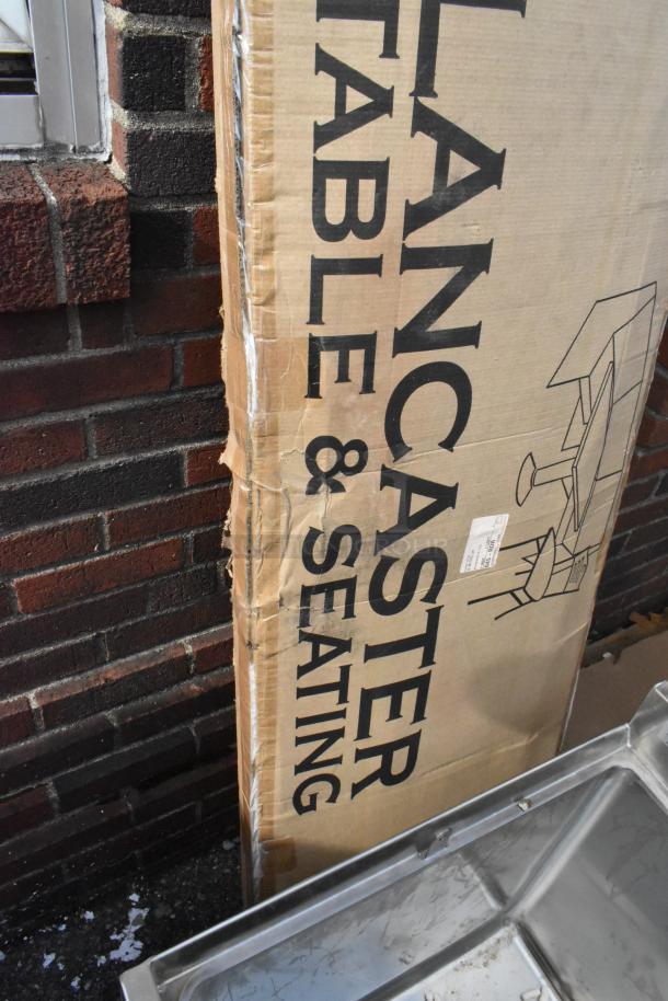 Boxed Lancaster Table & Seating 18"x72" folding table, brand new with visible scratches and dents on packaging.