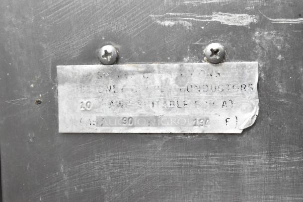 [label close-up, worn metal tag on stainless steel commercial brazing pan, faint text, secure screws]