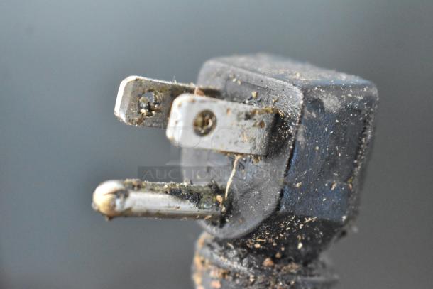 Close-up of a dirty, worn electrical plug with visible grime and minor rust. Two metal prongs are slightly bent.
