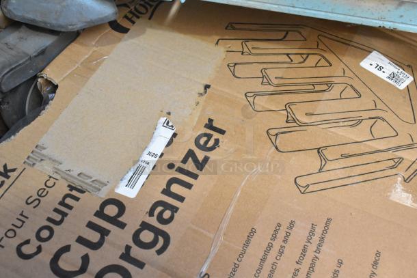 Box of a cup and lid organizer, with visible scratches and a torn label, indicating brand new scratch and dent condition.