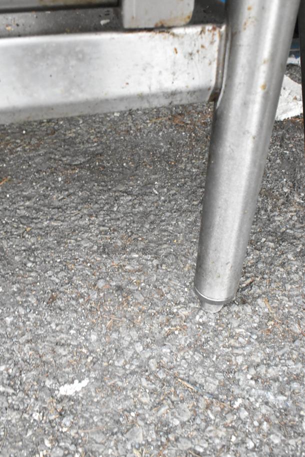 Stainless steel table leg on asphalt, showing part of under shelf and surface wear.
