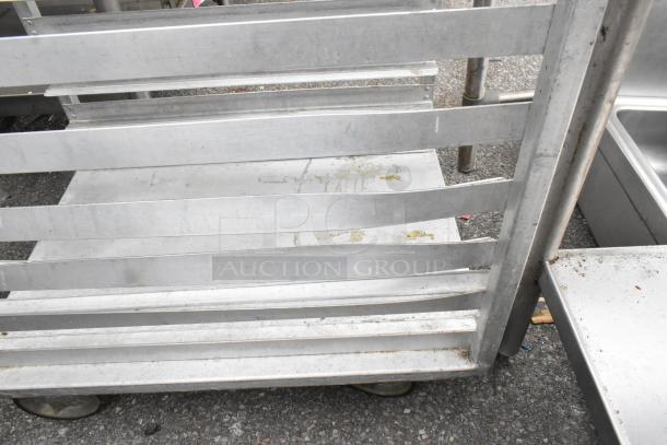 Metal pan transport rack on commercial casters. Features multiple shelves, visible wear, suitable for kitchen use.