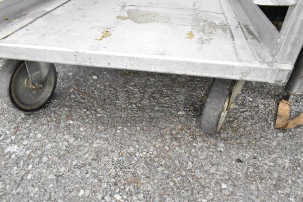 Metal pan transport rack on commercial casters, shows signs of wear, sturdy design. Suitable for kitchen or warehouse use.