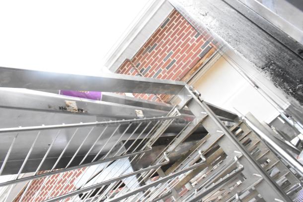 Metal commercial pan transport rack with wire shelves on commercial casters, visible wear, brick background.