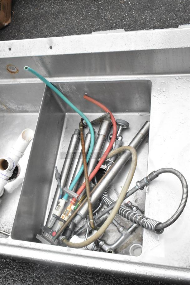 Stainless steel 3-bay sink with dual drain boards, seen without legs. Includes various plumbing attachments, indicating used condition.