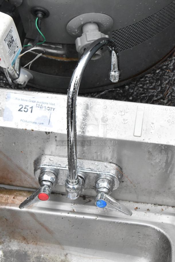 Stainless steel single bay wall mount sink with faucet, dual handles, and side splash guards. Some surface wear visible.