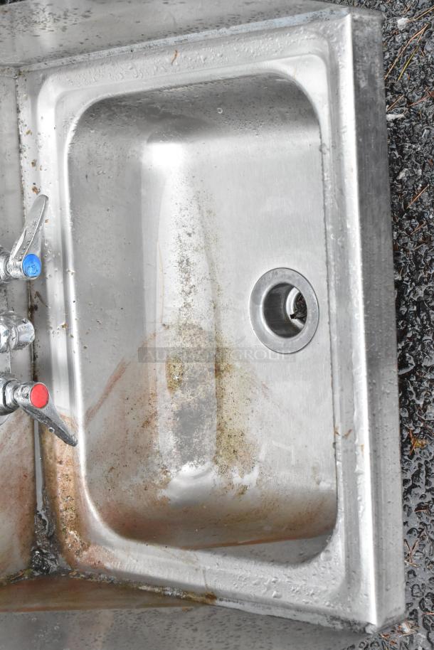 Stainless steel wall mount sink with faucet, handles, and side splash guards. Used condition with visible stains.