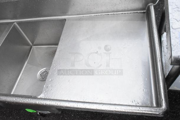 Stainless steel 3 bay sink with dual drain boards, no legs, visible drain, some surface wear and water droplets.