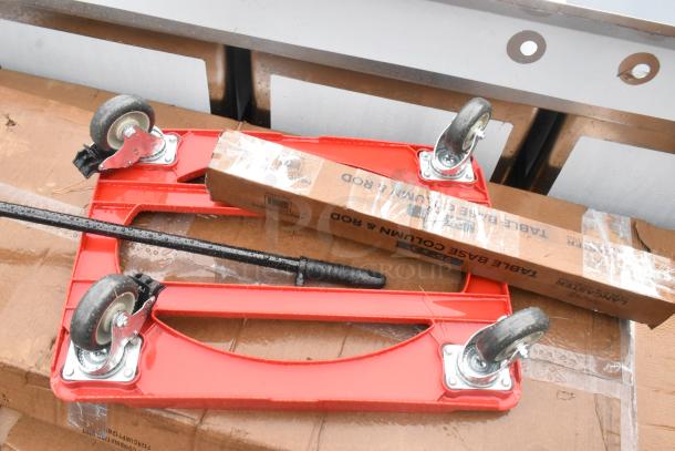 Red dolly with four caster wheels and boxed table base column and rod, new condition, on cardboard.