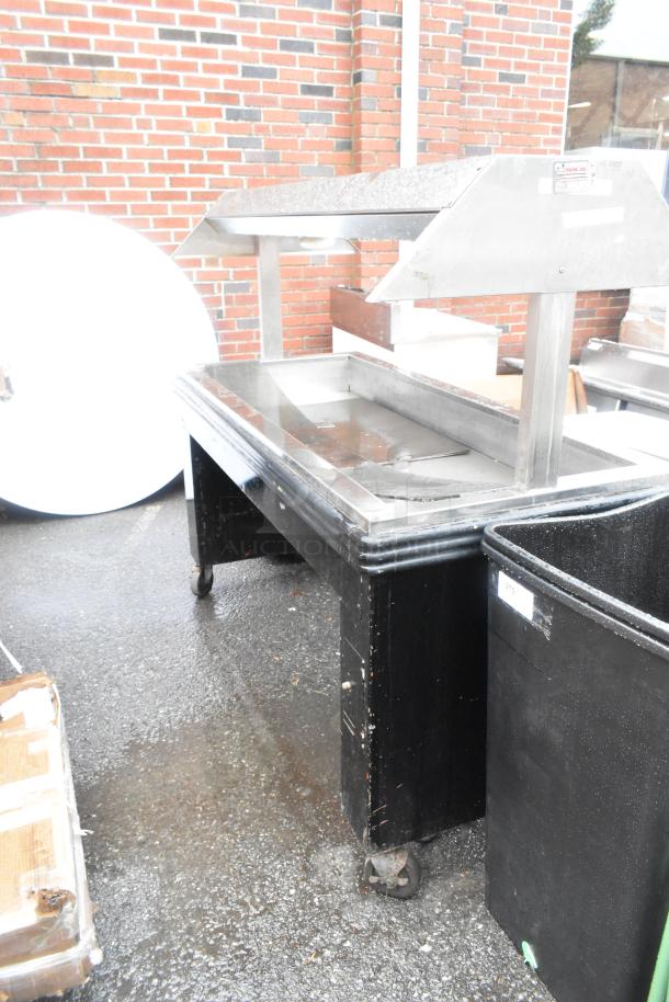 Metal commercial heated buffet station on casters, featuring a sleek stainless steel top with a built-in heat lamp. Visible wear on black base.