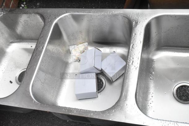 Stainless steel 3-bay drop-in sink with visible dents. Brand new, includes three boxed items in the left basin.