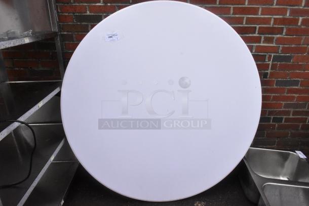 Brand new white round folding table with minor scratches and dents, visible label with item number, set against a brick wall.