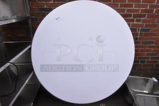 Brand new white round folding table with minor scratches and dents, visible label with item number, set against a brick wall.