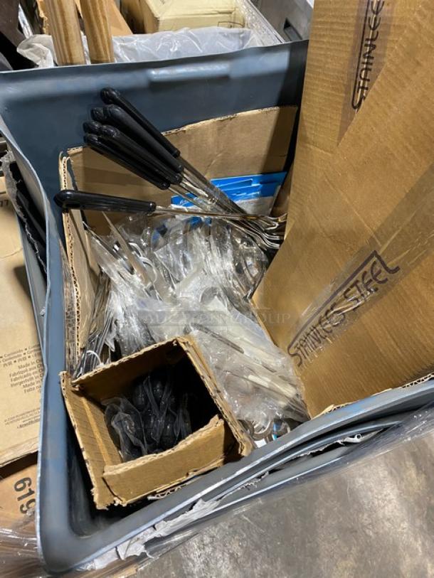 Box of assorted stainless steel kitchen utensils in a Rubbermaid trash bin. Black handles, cardboard packaging visible.