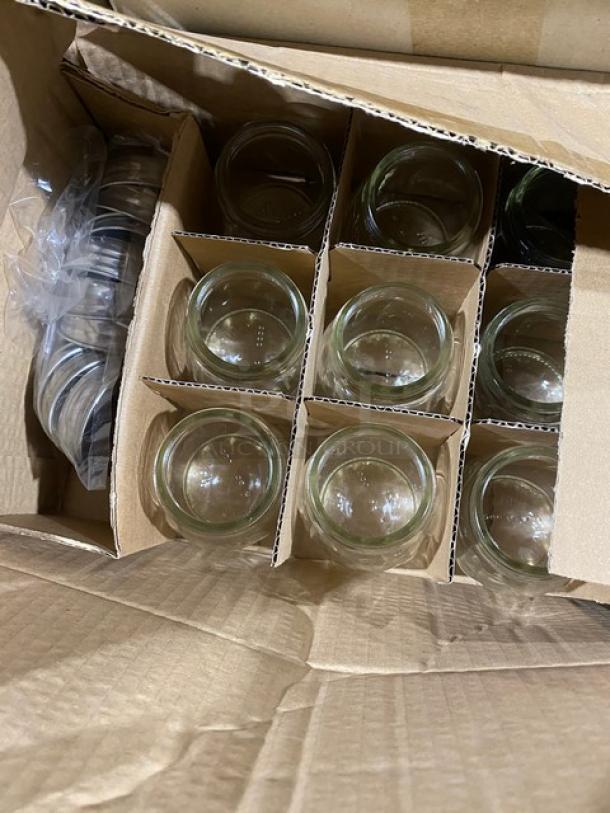 Box of clear glass cups with dividers, lids on the side, new condition in cardboard packaging.
