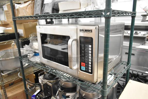 Brand new Solwave stainless steel commercial microwave oven, with visible scratches and dents, on a storage shelf.