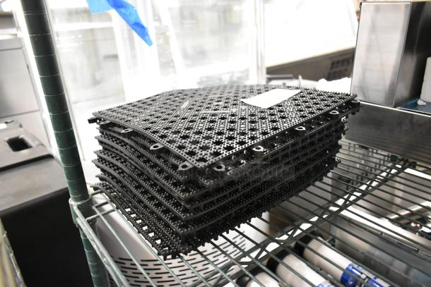 Stack of 10 brand new black floor mat pieces, labeled scratch and dent, on metal shelf, featuring textured surface.