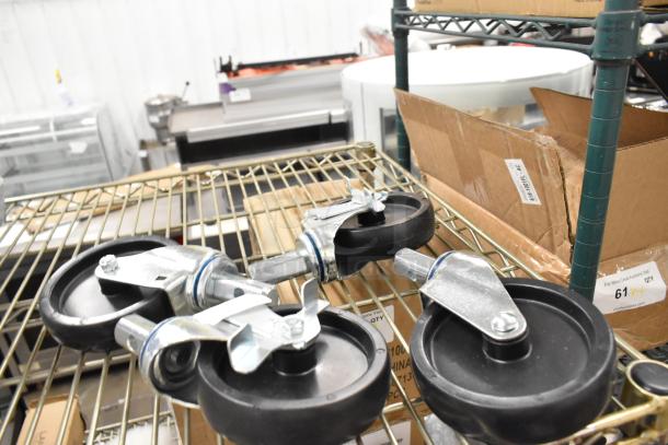 Lot of 4 brand new commercial casters with minor scratches and dents, black rubber wheels, metal mounts; shelf and boxes visible.