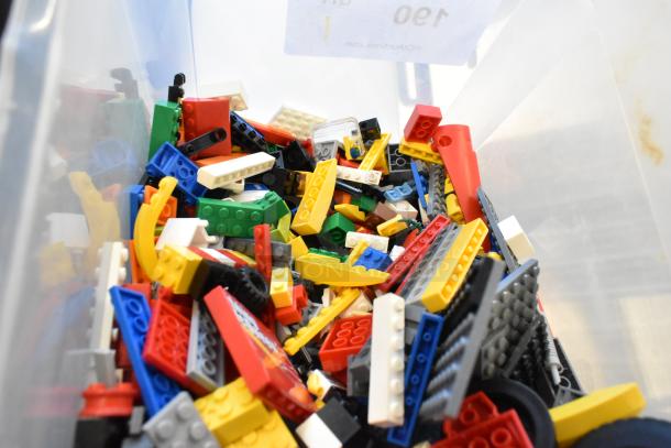 Box of assorted Lego pieces in various colors and shapes, includes blocks and wheels, in a clear plastic container.