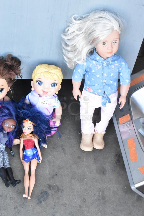 Lot of various dolls, including five with different hairstyles and outfits, in used condition on a concrete floor.